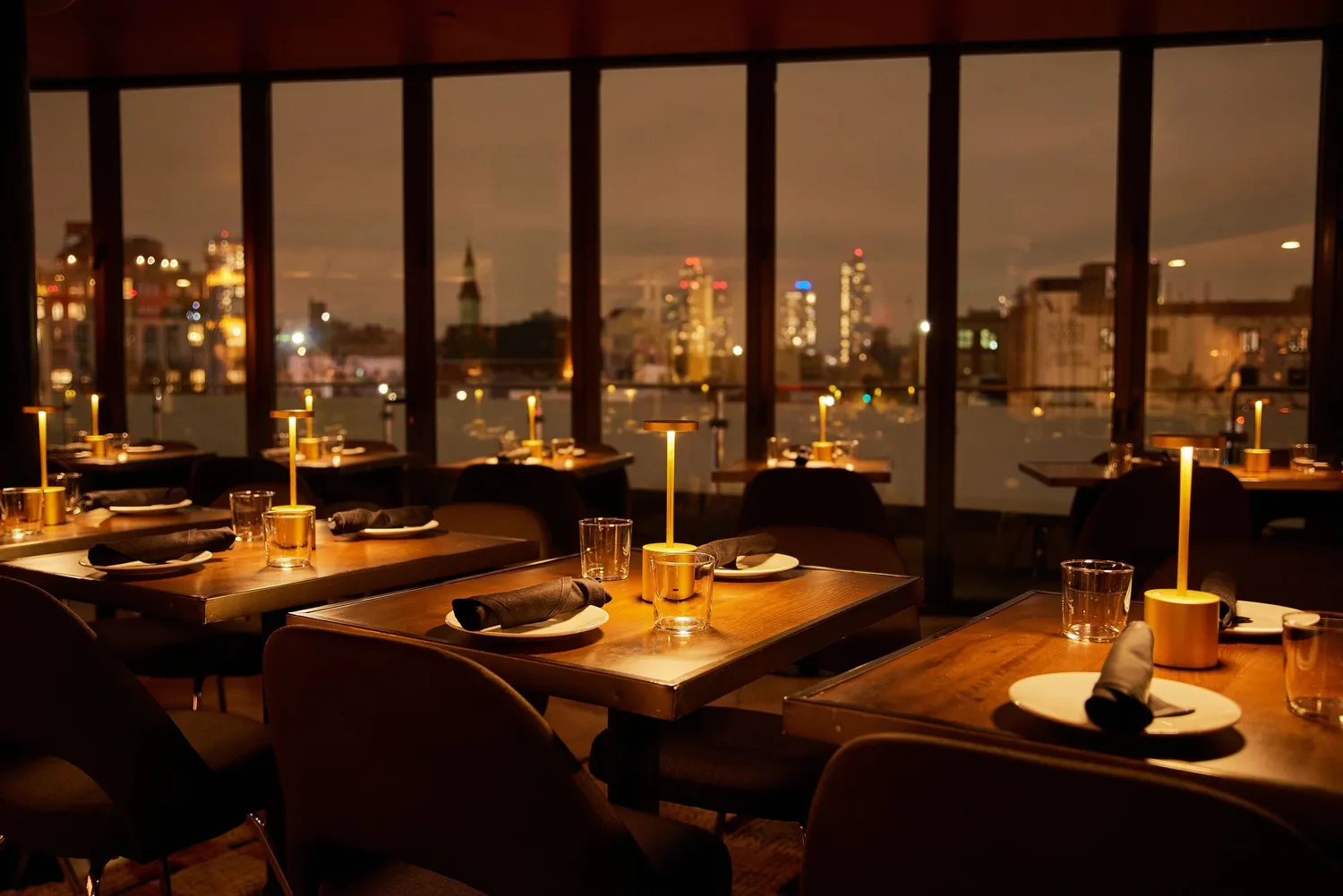 Cozy restaurant interior with wooden tables, elegant candles, and city skyline visible through large windows at night. Cozy restaurant interior with wooden tables, elegant candles, and city skyline visible through large windows at night.
