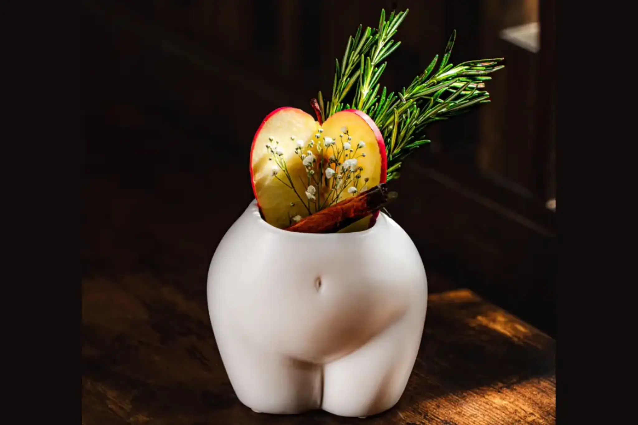 A quirky white vase shaped like a buttocks, filled with apple slices, sprigs of rosemary, and a stick of cinnamon on a wooden table.